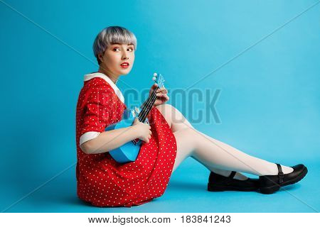 Close up portrait of beautiful dollish girl with short light violet hair wearing red dress holding ukulele over blue background. Copy space.