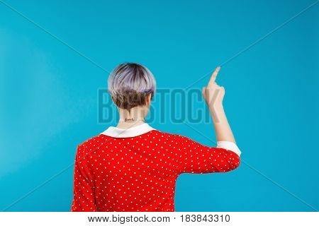 Close up portrait from back beautiful dollish girl with short light violet hair wearing red dress pointing with finger over blue background. Copy space.