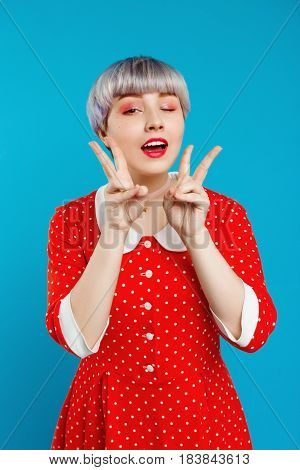 Close up portrait beautiful dollish girl with short light violet hair wearing red dresswinking showing victory gesture  over blue background. Copy space.
