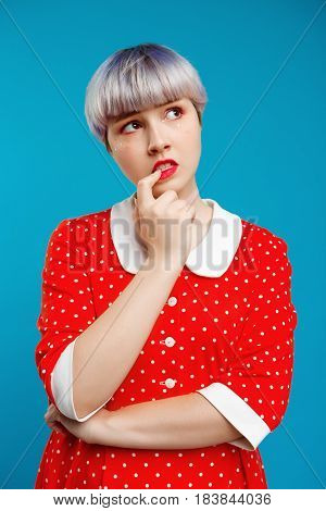 Close up portrait thoughtful beautiful dollish girl with short light violet hair wearing red dress over blue background. Copy space.