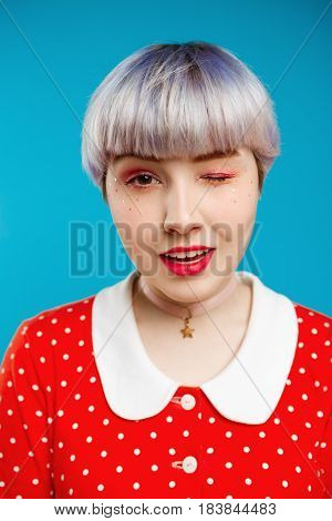 Close up portrait beautiful dollish girl with short light violet hair wearing red dress winking over blue background. Copy space.