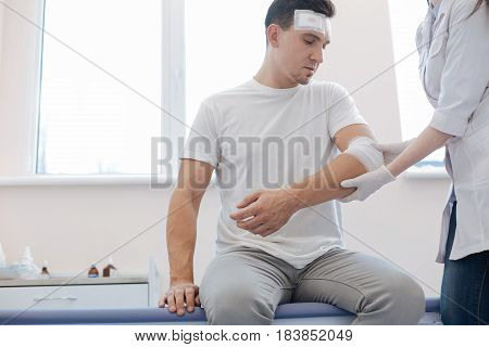Car accident. Cheerless wounded handsome man sitting on the medical bed and looking at his injury while being at the hospital