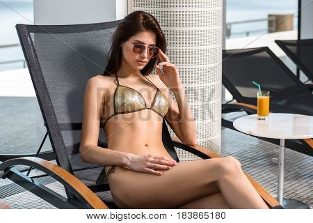 Young attractive slim girl in bikini relaxing on deck chair in wellness spa hotel resort. Near the table is a glass of orange juice