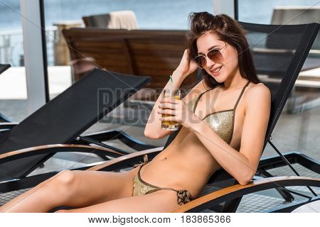Young attractive slim girl in bikini relaxing on deck chair in wellness spa hotel resort. Woman holding a glass of orange juice