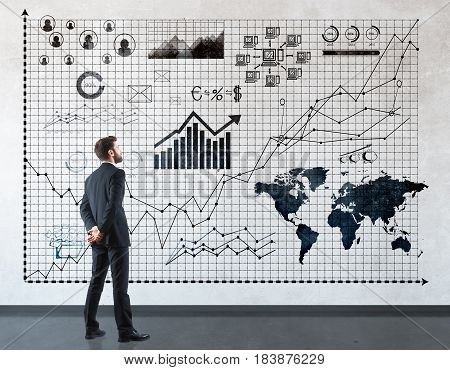 Back view of young businessman looking at business charts and diagrams in concrete interior. Finance concept