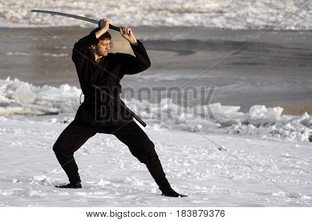 Ninja in black kimono with sword is practicing martial arts in the snow at winter.