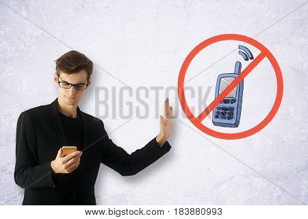 Young man on concrete background using smartphone despite the inhibitory sign. Addiction concept