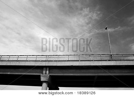 Street lamppost on transportation bridge under the cloudy sky as seen from below (black and white),