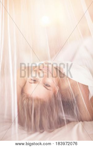 Joyful woman having fun in the bed, lying down behind transparent valance, enjoying romantic interior of bedroom, happy healthy lifestyle
