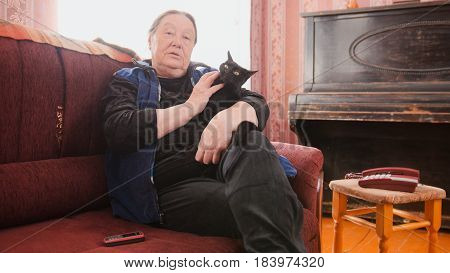 Portrait of old woman home - senior lady sits on sofa with black cat, telephoto