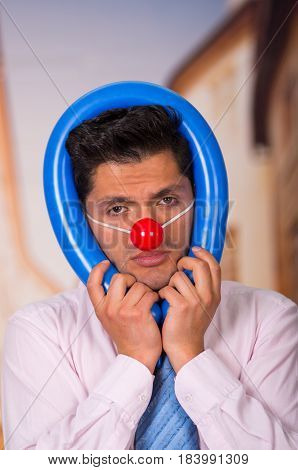 Unhappy handsome businessman with red plastic nose using a blue ballon around his head.