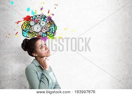Side view of an African American woman wearing a green shirt standing near a concrete wall with a colorful brains sketch and cogs on it. Mock up
