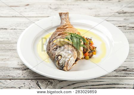 Dorada Stuffed With Vegetables. White Plate With Grilled Dorada Fish On A Wooden Table Close Up And 