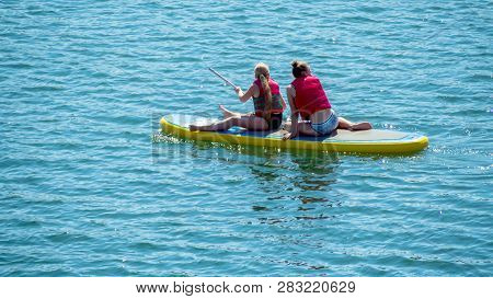 View Of 2 Young Girls Exercising With Sup Board In Turquoise Tropical Clear Waters. Sittiing And Pad