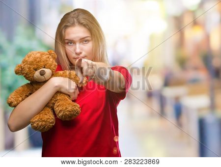 Young caucasian woman holding cute teddy bear over isolated background pointing with finger to the camera and to you, hand sign, positive and confident gesture from the front