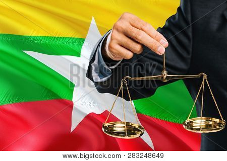 Burmese Judge Is Holding Golden Scales Of Justice With Myanmar Waving Flag Background. Equality Them
