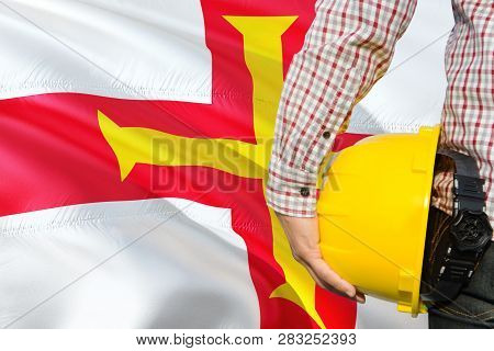 Engineer Is Holding Yellow Safety Helmet With Waving Guernsey Flag Background. Construction And Buil