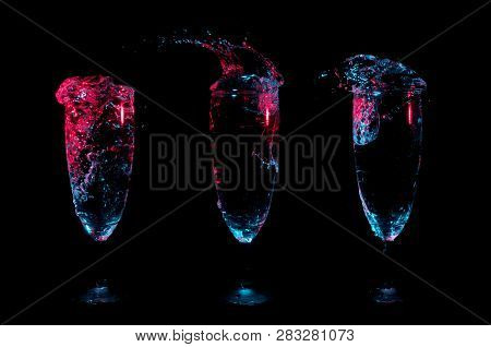 Three Glasses In A Row With Different Splashes Of Clear Beverages Under Red And Blue Lights On A Bla