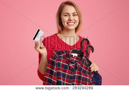 Positive European Lady With Toothy Smile, Holds Clothes On Hangers And Plastic Card, Pays For New Ou
