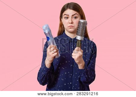 Studio Shot Of Puzzled Dark Haired Young Woman Holds Hairspray And Haircomb, Wears Fashionable Shirt
