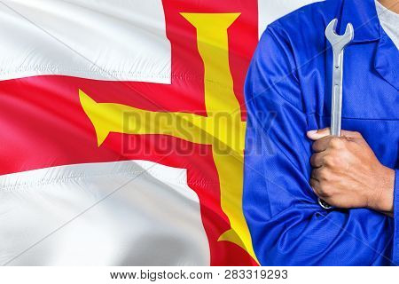 Mechanic In Blue Uniform Is Holding Wrench Against Waving Guernsey Flag Background. Crossed Arms Tec