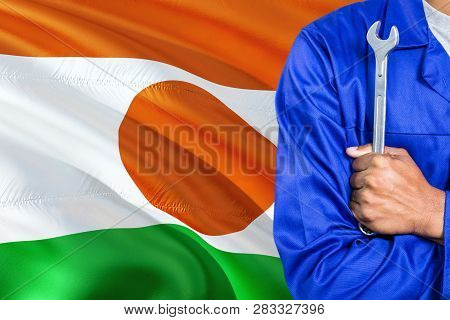 Nigerien Mechanic In Blue Uniform Is Holding Wrench Against Waving Niger Flag Background. Crossed Ar