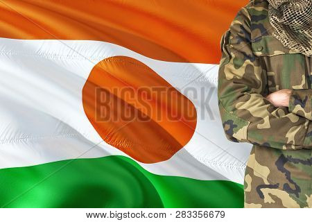 Crossed Arms Nigerien Soldier With National Waving Flag On Background - Niger Military Theme.