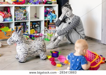 Tired Of Everyday Household Mother Sitting On Floor With Hands On Face. Kid Playing In Messy Room. S