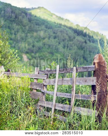 Weathered Wooden Split-rail Fence With Supports In Tall Grass With Mountains, Blue Sky And White Clo