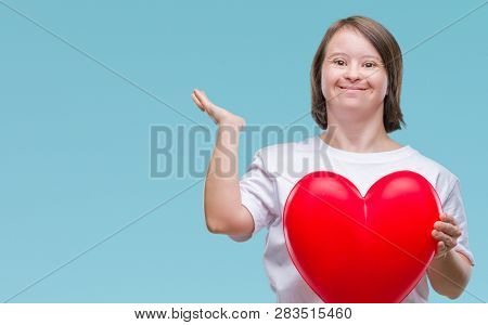 Young adult woman with down syndrome holding red heart over isolated background very happy and excited, winner expression celebrating victory screaming with big smile and raised hands