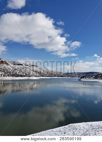 Snow Covered Hills, Trees, Clouds, And A Bright Blue Sky Reflect In The Partially Iced Over Prinevil