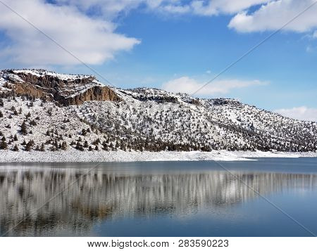 Snow Covered Hills, Trees, Clouds, And A Bright Blue Sky Reflect In The Partially Iced Over Prinevil