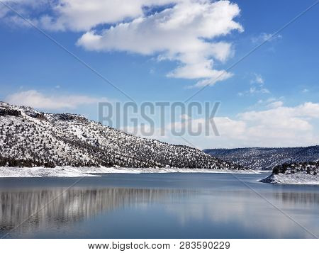 Snow Covered Hills, Trees, Clouds, And A Bright Blue Sky Reflect In The Partially Iced Over Prinevil