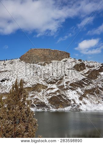 A Lone Juniper Tree In The Foreground Of An Basalt Outcropping On A Hill With A Fresh Layer Of Snow 