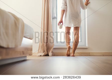 A Rear View Of Young Woman With Night Shirt Standing By The Window In The Morning.