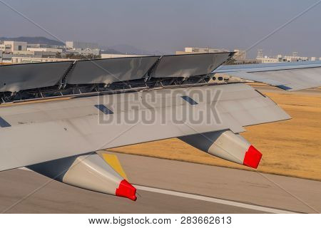 Aircraft Wing With Breaking Flaps Extended During Landing With Airport Buildings And Blue Sky In Bac