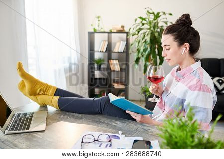 Side view portrait of relaxed young woman reading book and drinking wine with feet on desk, copy space