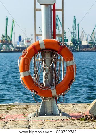 Red Orange Life Buoy Ring With Roap At The Edge Of The Pier Close-up. The  Seaport On A Sunny Day. W