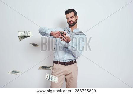 Finally Rich. Modern Looking Bearded Man Is Scattering Dollars While Standing On A Grey Background. 