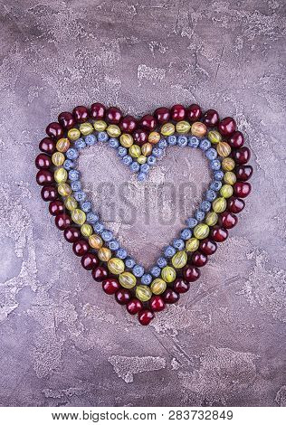 Fresh Sweet Cherries, Blueberry And Goosberry In Heart Shape Over On Concrete Gray Background. Top V