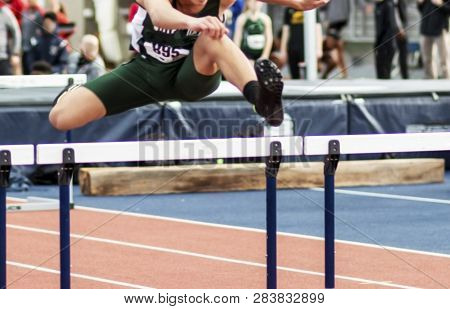 A High School Boy Is Running In A Hurdle Race In An Indoor Areana With People In The Background Watc
