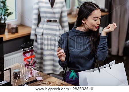 Dark-haired Smiling Asian Lady In A Dark-blue Blouse Choosing A Perfume