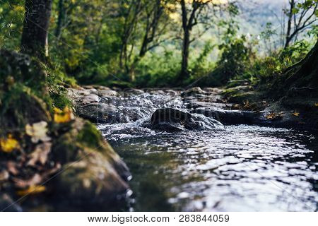 A River Creek With A Small Waterfall. Sunset And Strong Detail Bokeh View. Blue Calm Warm Water
