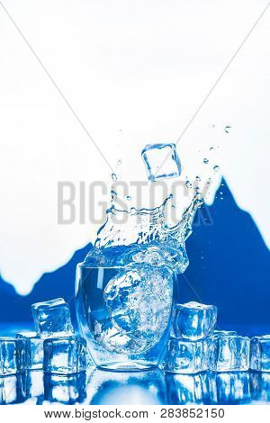 Crystal Clear Water Splash In A Double Wall Glass With Mountain Peaks On The Background. Ice Cubes I