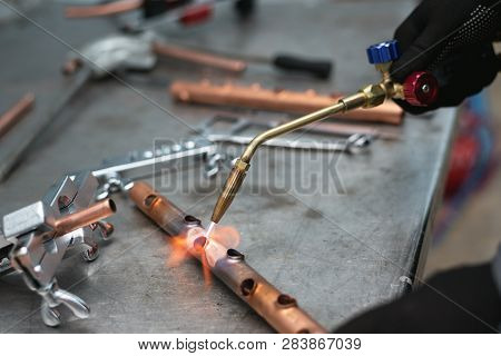 Worker Is Soldering A Pipe By A Blow Lamp On A Factory Workbench Background. Pipework.