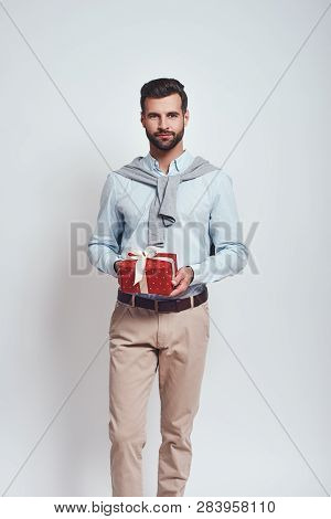 Present For You. Elegant Man In Casual Clothes Is Holding A Gift Box And Smiling At Camera While Sta