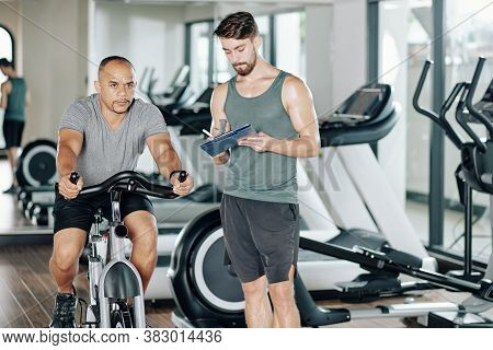 Fitness Trainer Writing In Document When His Client Trying New Cardio Workout And Riding Fast On Sta