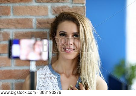 Beautiful Woman Holding A Smartphone At Selfie Stick. Young Female Smiles In Front Of Camera Over Br