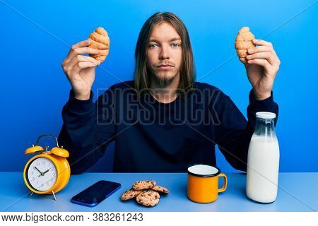 Handsome caucasian man with long hair eating croissant for breakfast relaxed with serious expression on face. simple and natural looking at the camera. 