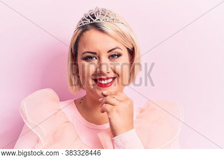 Beautiful blonde plus size woman wearing pincess tiara with diamonds over pink background smiling looking confident at the camera with crossed arms and hand on chin. Thinking positive.
